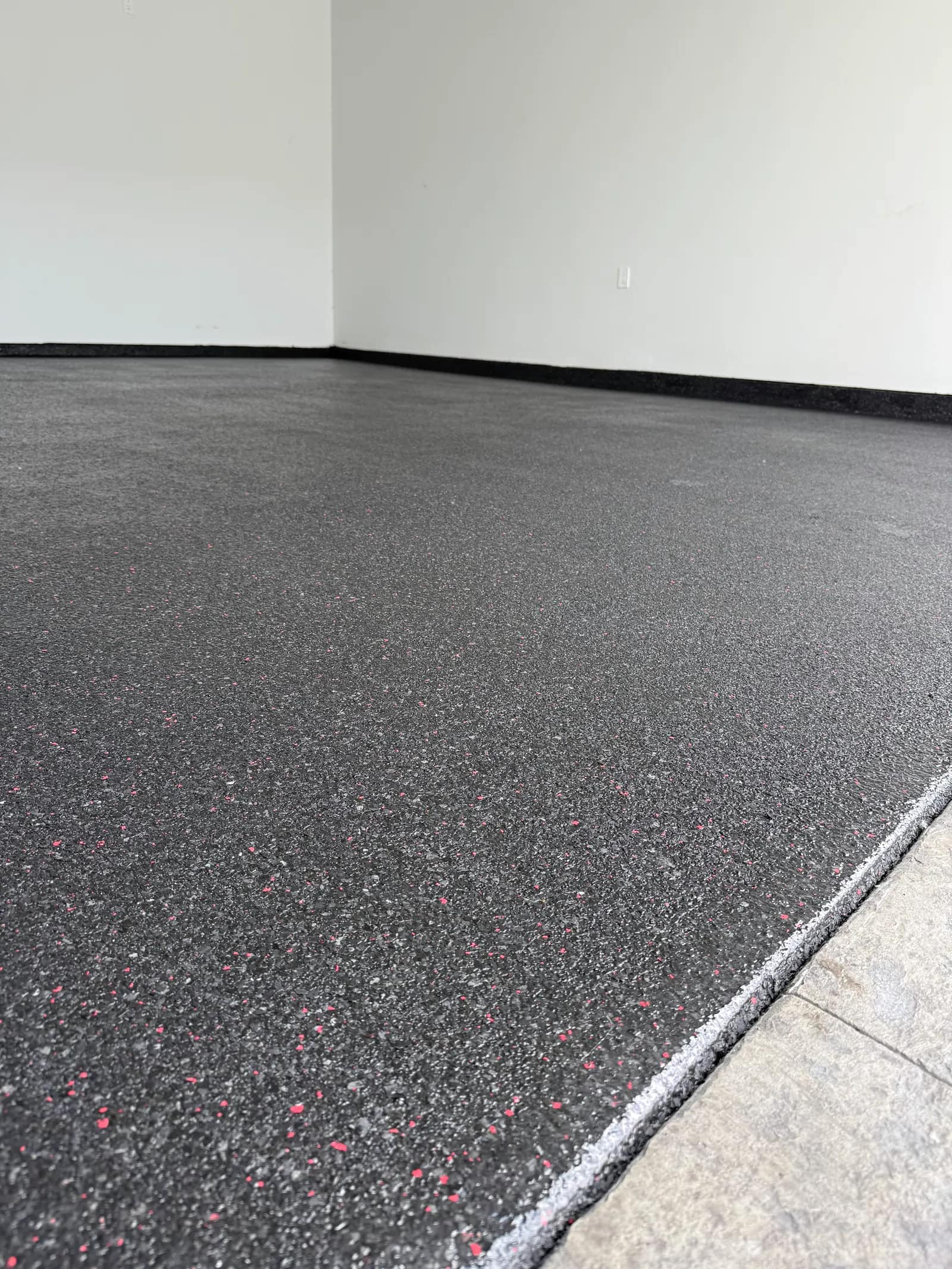 Matte black floor coating with fine red flecks, finished to a clean white edge at a tiled threshold